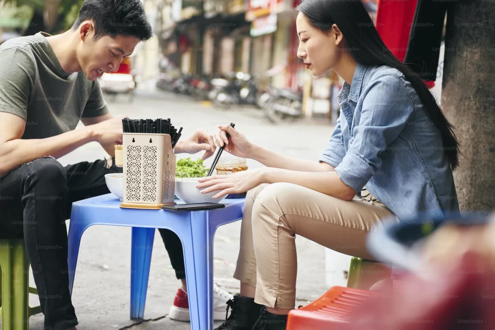 a man and a woman sat on small plastic stools, eating bowls of noodles in the street